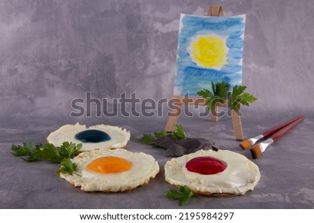 Artist supplies. Creative palette of colors made of fried eggs with yellow, red, blue yolk, painting brushes, easel with drawing and pieces of green parsley on gray concrete background