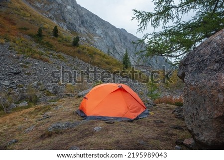 Dramatic mountain landscape with alone orange tent near big rock on hill with autumn flora against large mountain range in overcast. Lonely tent close-up among fading autumn colors in high mountains.