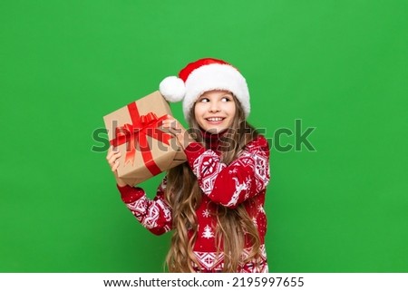 A little girl holds a gift on a green isolated background. A child in a Santa Claus hat and a warm sweater holds a gift box.
