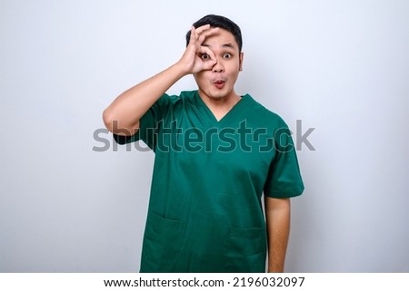Amused happy, handsome asian male doctor, intern in scrubs looking through hand binoculars with excited expression, smiling over white background