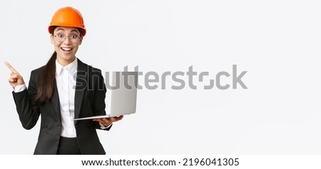 Excited happy asian female engineer, industrial woman in safety helmet and business suit, showing presentation, pointing finger at graph or chart and holding laptop computer, smiling amazed