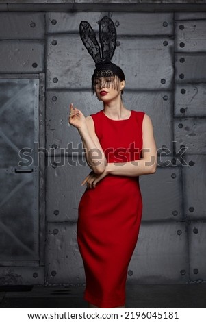 Young woman in black rabbit or hare fancy mask and red dress. In the background there is a metal wall with rivets .