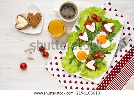 Breakfast on Valentines Day. A plate of fried heart-shaped eggs, cheese and tomatoes, coffee and juice, bread and butter on a white wooden table and the inscription I love you Breakfast for loved
