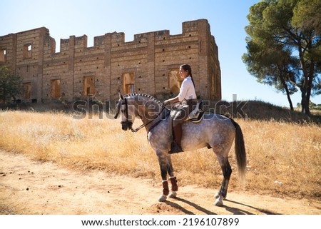 Young and beautiful Spanish woman on a Thoroughbred horse riding in the countryside in Spain. The woman is wearing a horse riding uniform. Thoroughbred and equine concept. Royalty-Free Stock Photo #2196107899