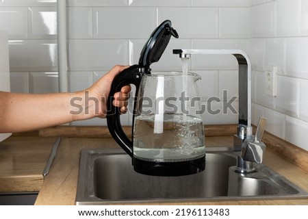 A woman draws water into an electric kettle in the background of the kitchen. It's time for breakfast and tea. Modern electric kettle on a wooden table. Kettle for boiling water. Royalty-Free Stock Photo #2196113483