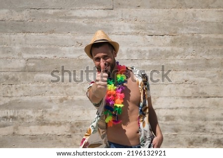Young and handsome man, blue eyes, with beard, hat, open Hawaiian shirt and flower necklace and making ok sign with thumb on a gray background. Concept holidays, party, trips. Selective focus.