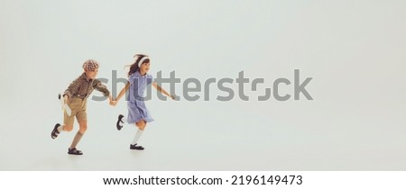 Portrait of stylish children, boy and girl, holding hands, running, posing isolated over grey studio background. Concept of childhood, friendship, fun, lifestyle, fashion, retro style