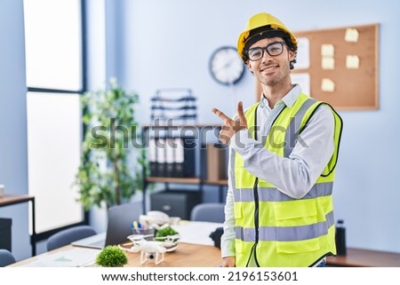 Hispanic man wearing architect hardhat smiling cheerful pointing with hand and finger up to the side 
