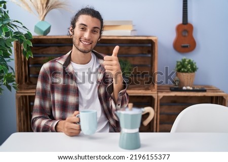 Young hispanic man drinking coffee from french coffee maker smiling happy and positive, thumb up doing excellent and approval sign 