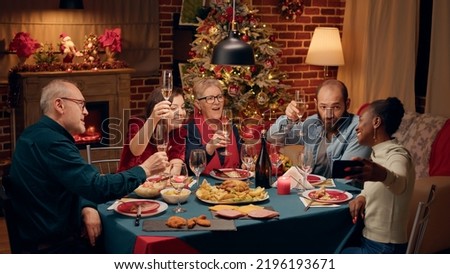 Beautiful african american woman with smartphone taking selfie photo of family celebrating winter holiday at home. Joyful diverse people enjoying Christmas dinner while creating memories.