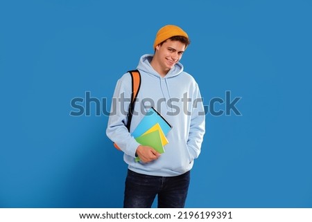 Photo of young fashionable student guy with cap and backpack smiling at camera, holding notebooks in hand, isolated over blue studio background. A lot of copy space. 
