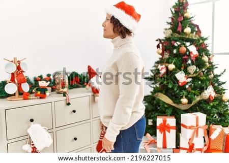 Middle age hispanic woman standing by christmas tree looking to side, relax profile pose with natural face with confident smile. 