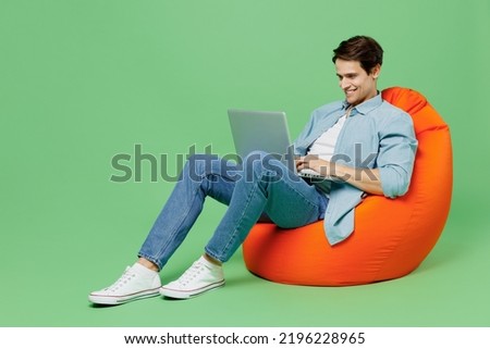 Full size body length young brunet man 20s wears blue shirt sit in bag chair hold use work on laptop pc computer typing browsing chatting send sms isolated on plain green background studio portrait