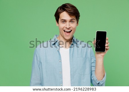 Shocked happy surprised overjoyed young brunet man 20s years old wears blue shirt hold in hand use mobile cell phone with blank screen workspace area isolated on plain green background studio portrait