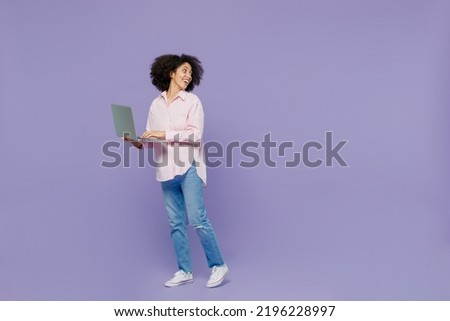 Full body fun young woman of African American ethnicity 20s wear pink striped shirt hold use work on laptop pc computer look aside on workspace isolated on plain pastel light purple background studio
