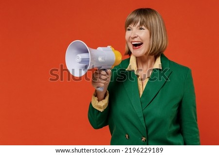 Elderly woman 50s wearing green classic suit hold scream in megaphone announces discounts sale Hurry up isolated on plain orange color background studio portrait. People business lifestyle concept.