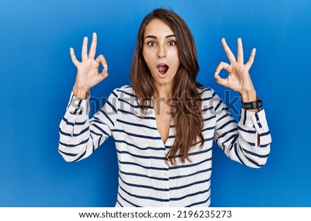 Young hispanic woman standing over blue isolated background looking surprised and shocked doing ok approval symbol with fingers.