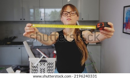 Indoor portrait of confident focused red-haired woman with tape measure in hands showing it to the camera while standing on background of workshop. High quality photo