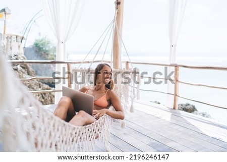 Full length of positive young self employed woman in stylish bikini smiling and admiring ocean while lying in hammock and working remotely on laptop during summer holidays