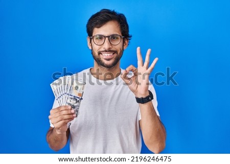 Handsome latin man holding dollars banknotes doing ok sign with fingers, smiling friendly gesturing excellent symbol 