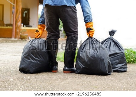 Closeup man holds black plastic bag that contains garbage inside, stand in front of house. Concept : Waste management. Environment problems. Daily chores. Throw away rubbish .                          Royalty-Free Stock Photo #2196256485