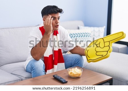 Young hispanic man supporting soccer team sitting on sofa at home