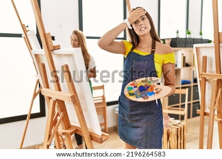 Young hispanic woman at art classroom stressed and frustrated with hand on head, surprised and angry face 