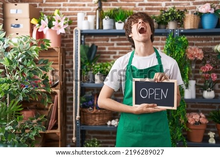 Hispanic young man working at florist holding open sign angry and mad screaming frustrated and furious, shouting with anger looking up. 