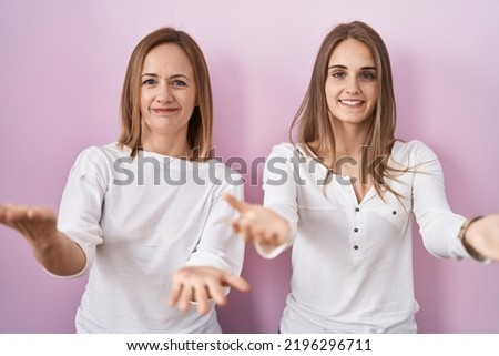 Middle age mother and young daughter standing over pink background smiling cheerful offering hands giving assistance and acceptance. 
