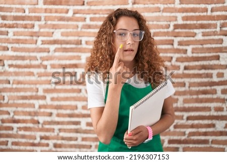 Young caucasian woman holding art notebook pointing to the eye watching you gesture, suspicious expression 