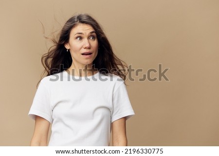 an emotional, funny woman with long, well-groomed hair blowing in the wind is standing in a white t-shirt with a very frightened expression on her face. Horizontal photo on a beige background