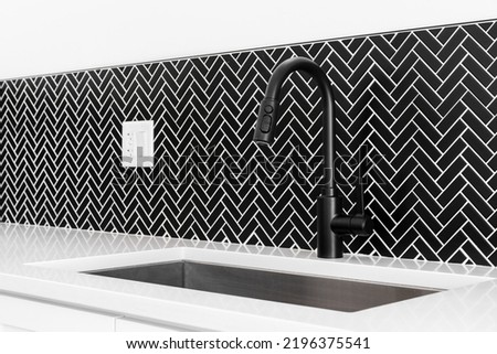 A kitchen sink detail shot with a black faucet, black marble herringbone backsplash, and a white granite countertop. Royalty-Free Stock Photo #2196375541