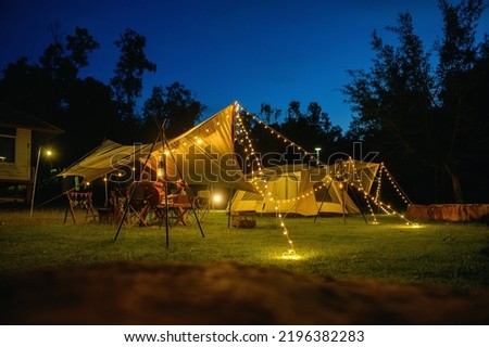 Outdoor camping tent with tarp or flysheet on grass courtyard and warm night light under dark blue sky twilight time, family vacation picnic on holiday relax, Overview of camping of family tourist. Royalty-Free Stock Photo #2196382283