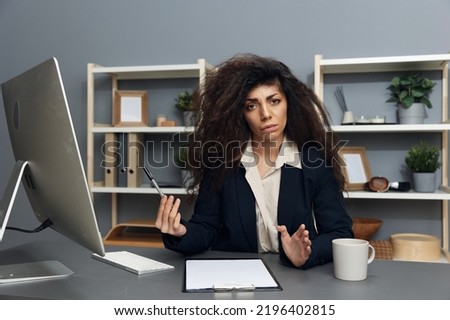 Confused angry tanned adorable curly Latin businesswoman in jacket hold hand up irritated looks at camera in office interior. Copy space Banner. Corporation leader lady uses modern computer in work