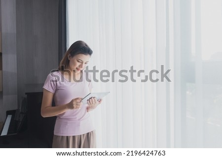 Asian businessman works from home using a tablet device, browsing the Internet, playing social media, Attractive businesswoman using digital tablet while standing in front of a window in home