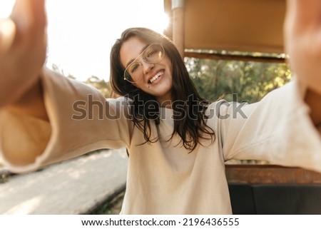 Smiling young caucasian girl takes selfie stretching hands to camera in sunny weather outdoors. Brunette wear glasses, everyday sweatshirt. Emotion concept