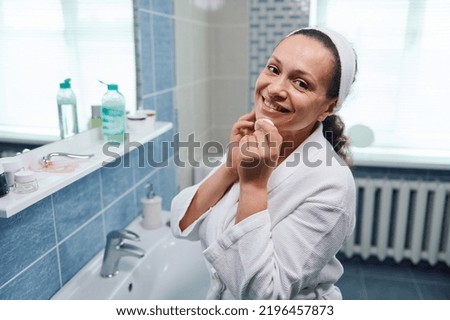 Middle-aged dark-haired charming woman in white bathrobe, using a face cleanser and a cotton pad, removes make-up, and cutely smiles looking at camera standing in the home bathroom. Skin and body Royalty-Free Stock Photo #2196457873