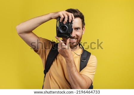 Happy male tourist with backpack taking picture on professional DSLR camera over yellow studio background. Caucasian man shooting travel photos during his adventures