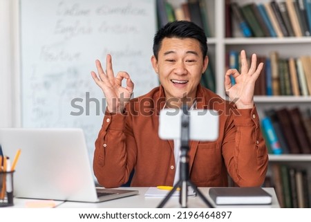 E-Teaching Is Okay. Cheerful Japanese Male Tutor Having Online Class Via Laptop And Smartphone And Gesturing OK Sitting At Workplace Indoor. Teacher Approving Distance Education. Selective Focus