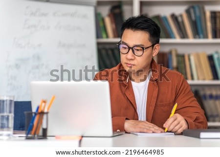 E-Teaching. Korean Middle Aged Man Teacher Using Laptop And Taking Notes Preparing For Virtual Lecture Sitting At Desk In Modern Classroom. Internet Technology And Education. Selective Focus