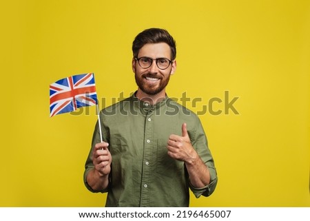 Happy man holding flag of UK and showing thumb up, recommending online educational course, learning English on Internet, posing over yellow background Royalty-Free Stock Photo #2196465007
