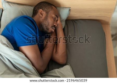 High Angle Shot Of Sleeping Black Guy Napping Holding Hands Near Face In Bed At Home. Male Resting In Modern Bedroom Enjoying Healthy Sleep. View From Above. Recreation Concept