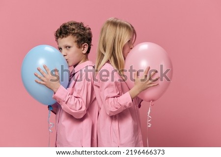 cute happy kids in pink clothes on a pink background stand sideways to the camera and the boy kisses his blue balloon. Horizontal studio photography with empty space