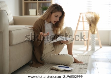Shocked woman reading letter while sitting on floor near sofa at home Royalty-Free Stock Photo #2196502999