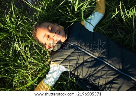 Upper view of happy carefree black boy kid having rest after active outdoor games lying on green juicy grass in city park, enjoying warm water, blue sky and tranquility of moment. Happy childhood Royalty-Free Stock Photo #2196505571