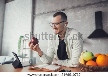 A caucasian man eating an apple looking into the tablet at the table early in the morning in a home kitchen. Business people communication. Kitchen interior