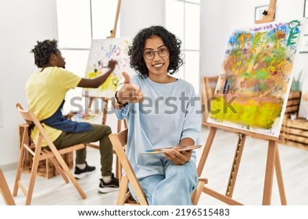 Young hispanic woman at art studio smiling cheerful offering palm hand giving assistance and acceptance. 