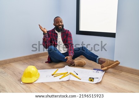African american man sitting on the floor at new home looking at blueprints smiling cheerful presenting and pointing with palm of hand looking at the camera. 