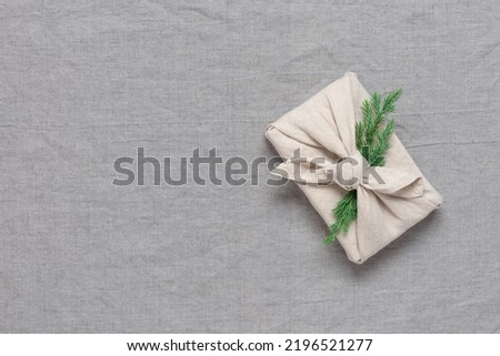 A gift wrapped in a fabric with a coniferous green branch, gray linen background. Gift in furoshiki style. Christmas and zero waste concept. View from above.