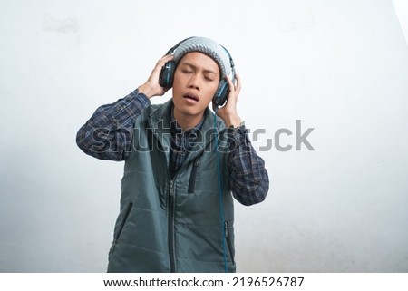 asian young man wearing dark blue vest and beanie wearing headphones, isolated on white background listening to music leisurely.